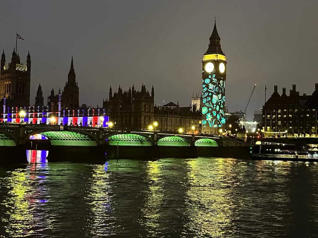 An evening view of Big Ben and the Palace of Westminster taken across the water. Red, white and blue light displays are projected on the front of Westminster, and blue flowers are projected on Big Ben.