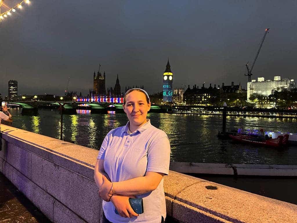 A young person stands smiling next to a low stone wall. In the background is the River Thames and beyond that, Big Ben and the Palace of Westminster lit with red, white and blue light displays.
