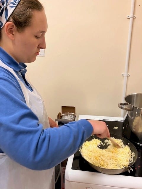 young person in a blue jumper and while apron stirring a pan filled with grated cheese and other ingredients.